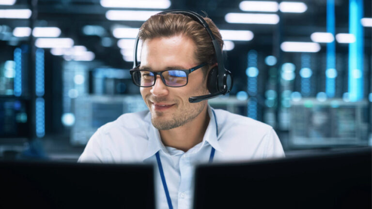 Man in business shirt wearing a headset sitting in front of two computer monitors.